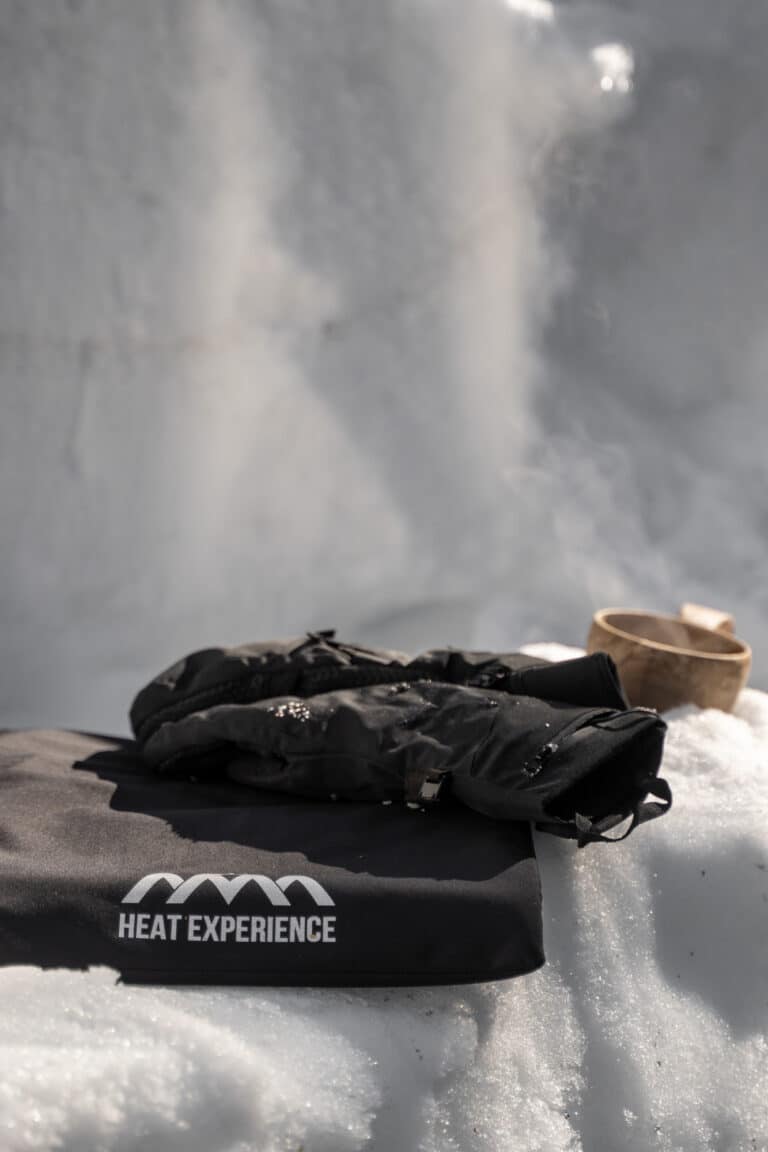 Schwarze Handschuhe mit "Heat Experience"-Logo auf Schnee, eine Holzschale im Hintergrund, Dunst steigt auf.
