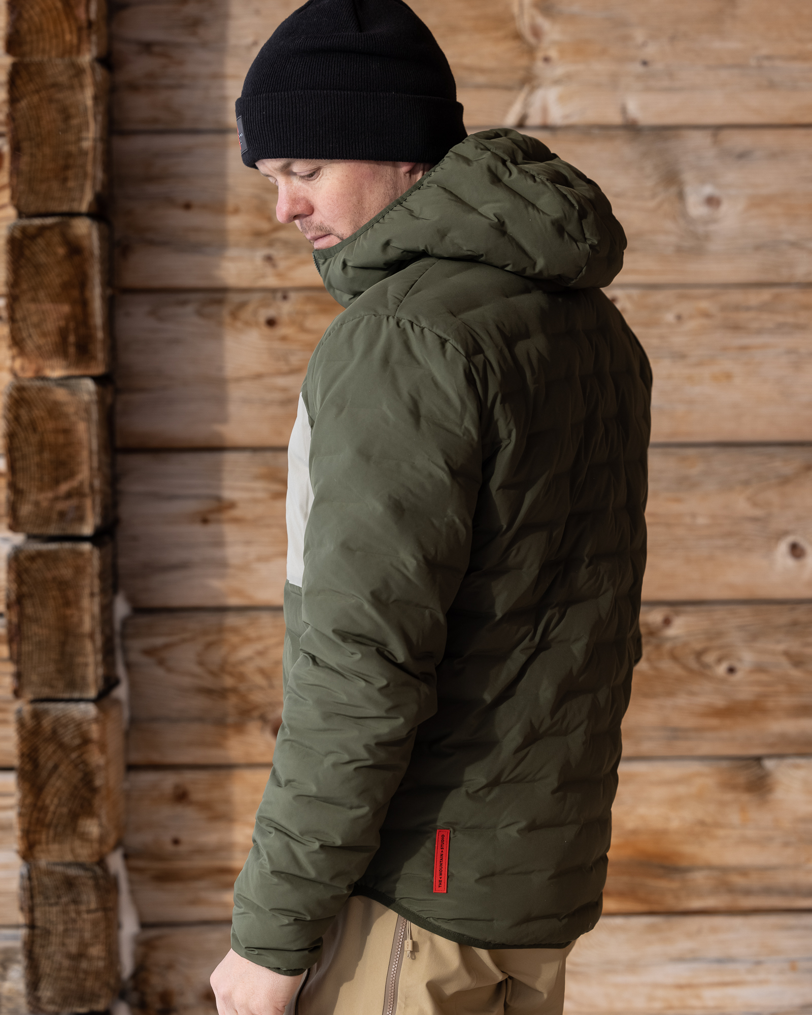Ein Mann mit Mütze trägt eine wendbare, hellgrüne D-3L Kapuzenjacke vor einer Holzwand.
