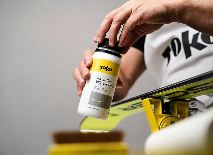 Une main ouvre un flacon de "TOKO All-in-One Wipe & Brush" au-dessus d'un ski. Le ski est posé sur un dispositif jaune.