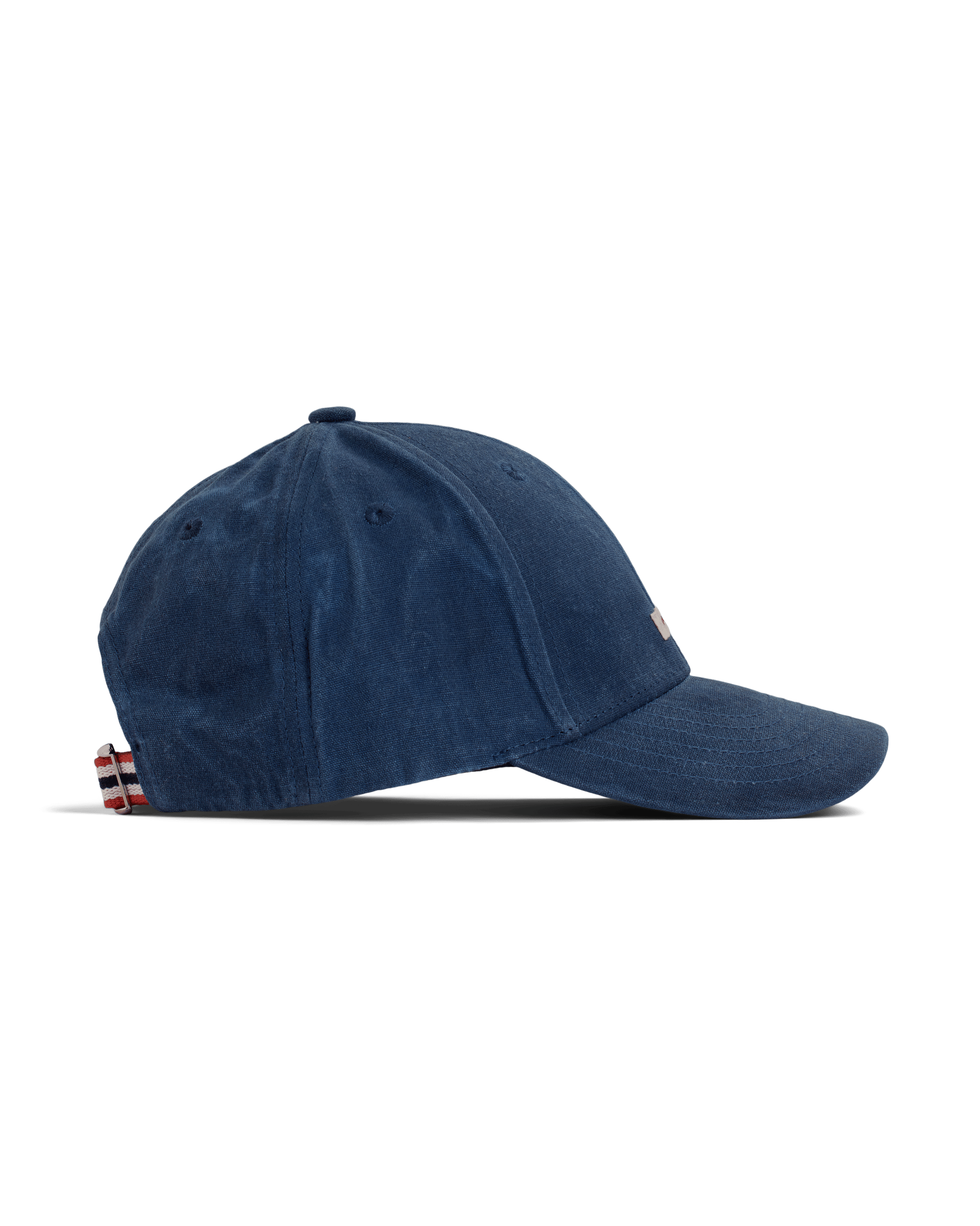 Seitliche Ansicht einer marineblauen Amundsen Waxed Cotton Cap vor einem weißen Hintergrund. Die Kappe hat einen Knopf oben, einen gebogenen Schirm und einen Riemen mit rot-weiß-blauen Streifen am Hinterkopf zur Größenverstellung.