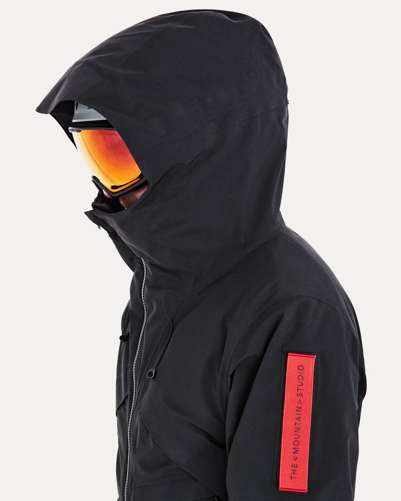 Person mit Skibrille und Kapuze einer schwarzen Gore-Tex 2L Stretch Insulated Jacke. Am Ärmel ein rotes Label.