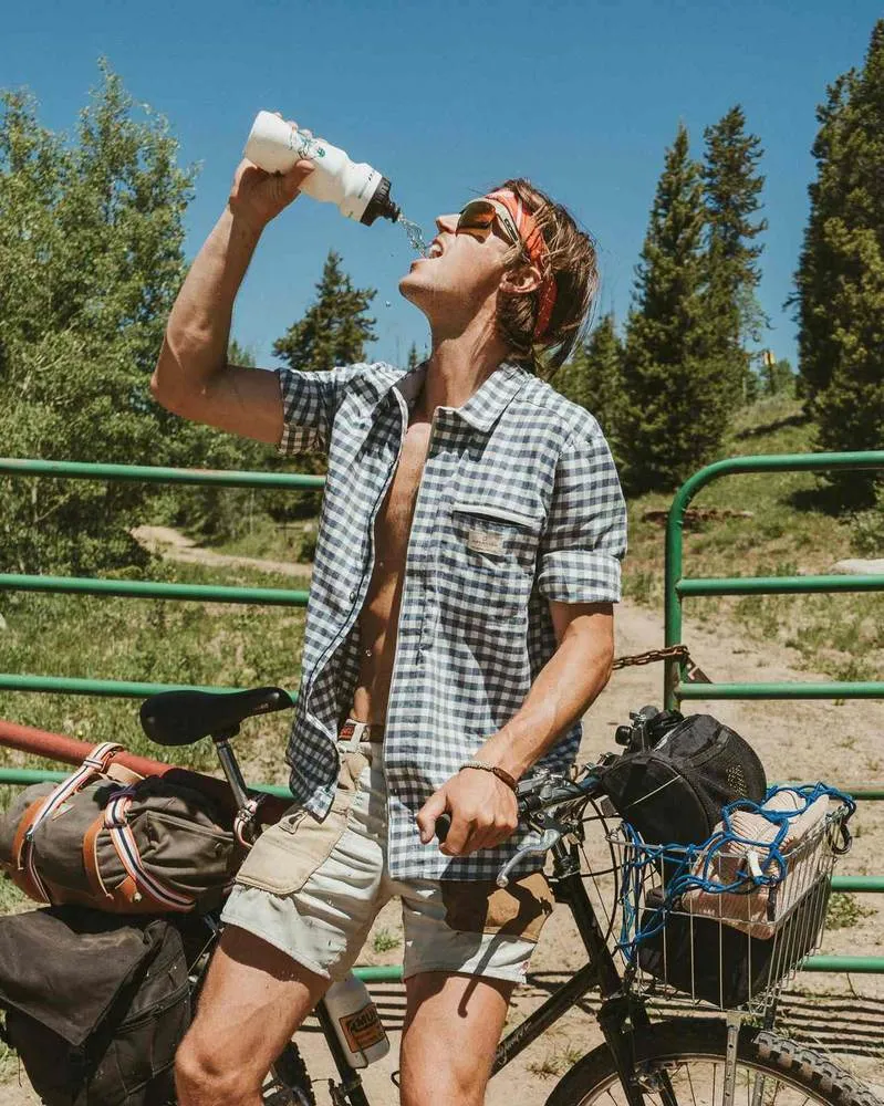 A man in a plaid shirt and shorts drinks water from a bottle, leaning against a bicycle in front of a green barred door.