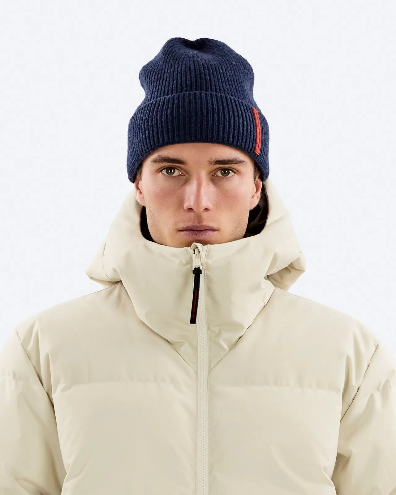 Ritratto di un uomo con berretto blu e piumino beige davanti a uno sfondo bianco.