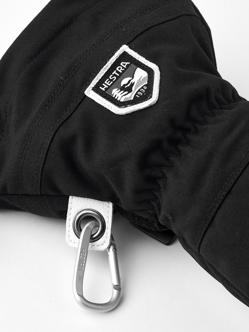Detailaufnahme eines schwarzen Handschuhs mit Hestra-Logo, weißer Schlaufe und Karabinerhaken.