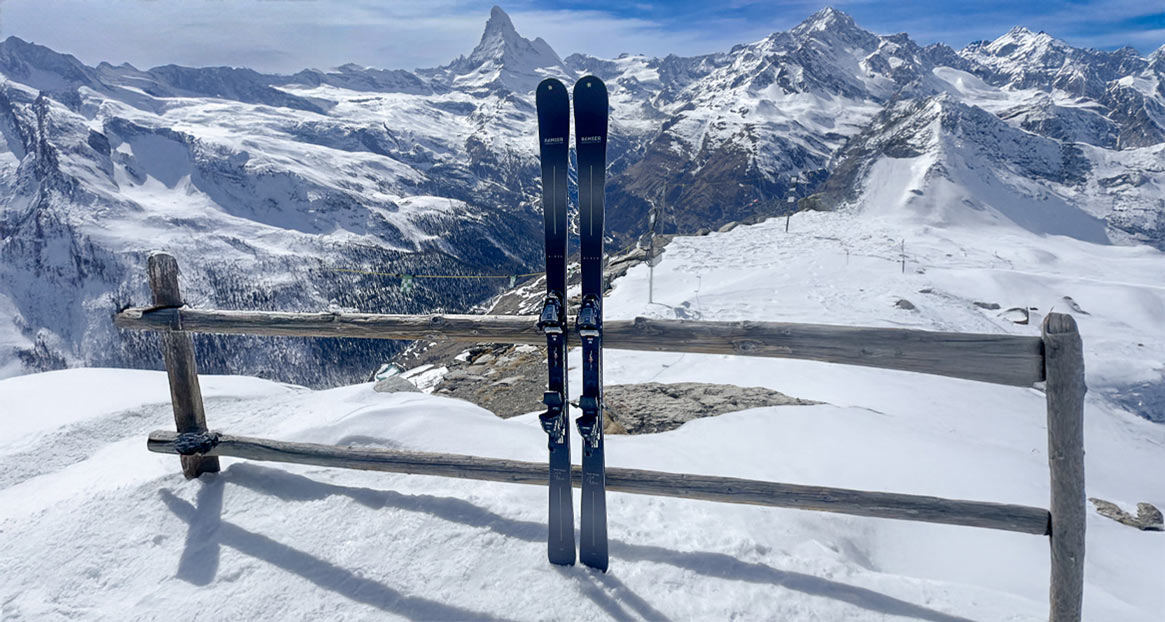 Zwei Skier stehen an einem Holzzaun im Schnee, im Hintergrund sind schneebedeckte Berge unter einem blauen Himmel zu sehen.