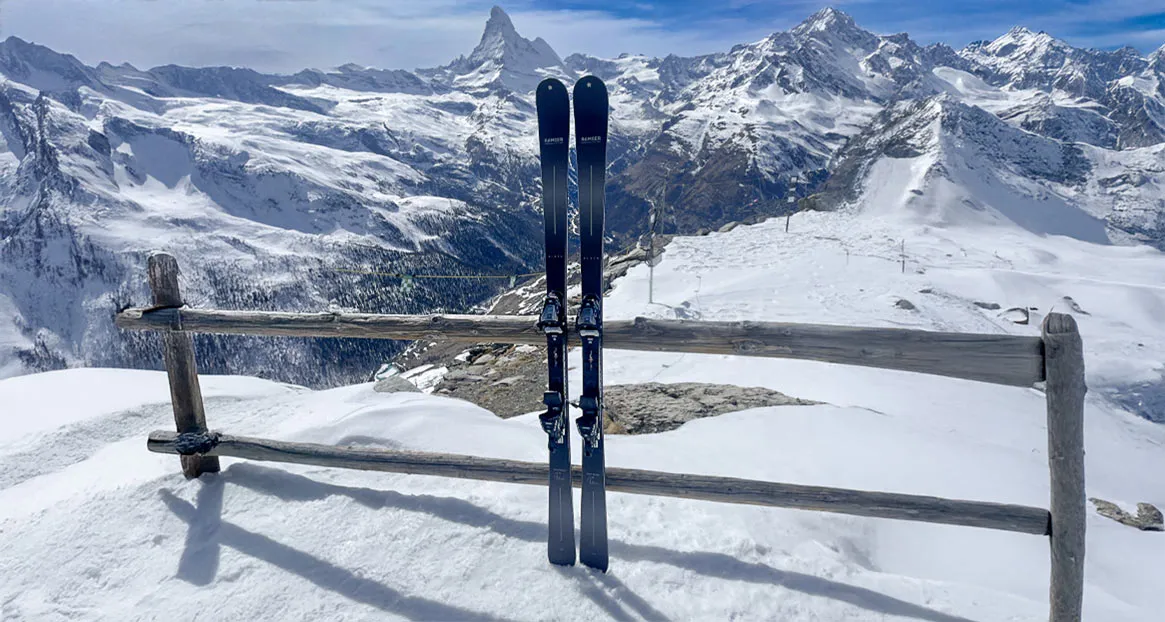 Zwei Skier stehen an einem Holzzaun im Schnee, im Hintergrund sind schneebedeckte Berge unter einem blauen Himmel zu sehen.