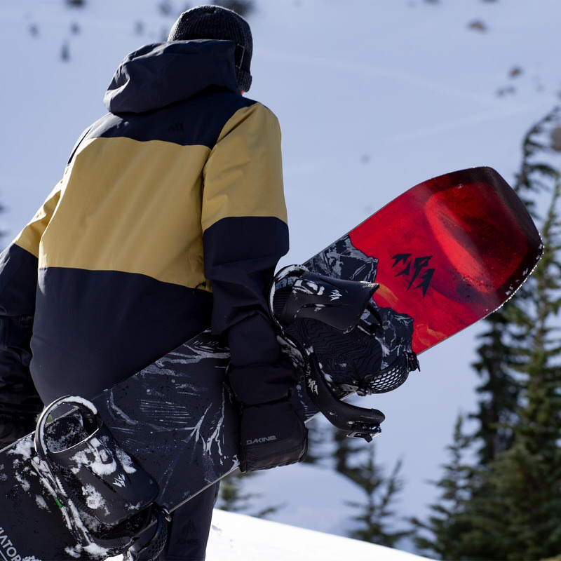 Person mit Snowboard Jones Aviator 2.0 LTD, rote und schwarze Grafik. Schnee liegt auf dem Board, Bindungen montiert. Winterkleidung.