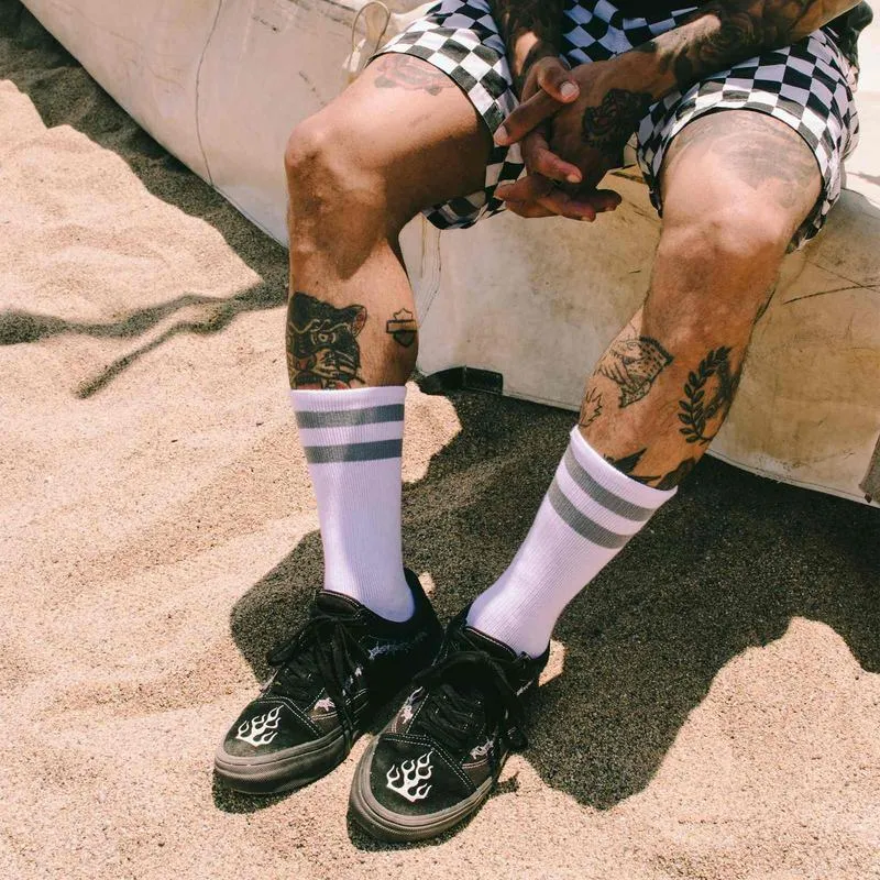 Person mit Tattoos trägt Schachbrettmuster-Shorts, Falkor Mid High Socken, und Turnschuhe am Strand.