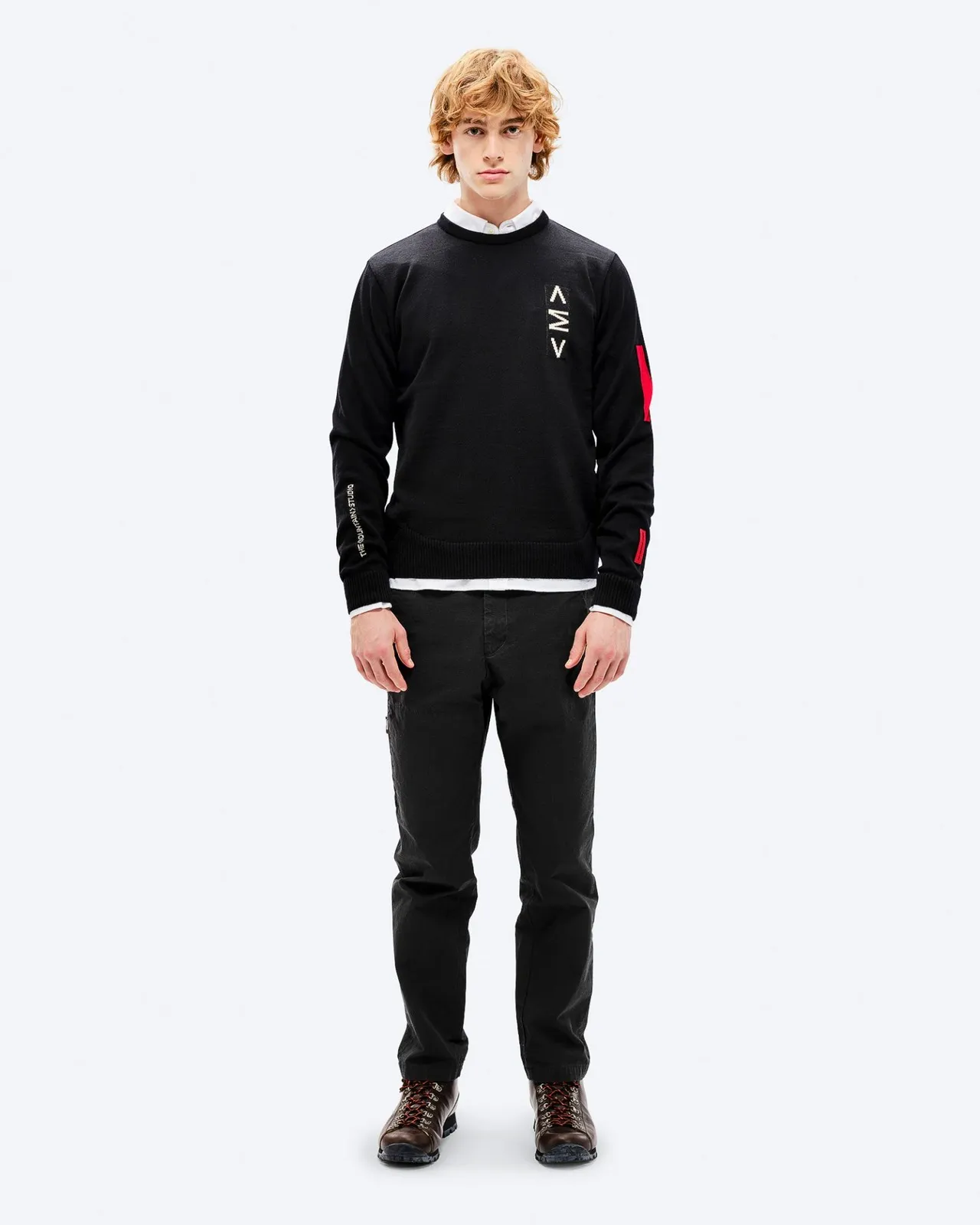Mann mit rotem Haar trägt schwarzen K-3 Ski Sweater mit roten Akzenten und schwarzen Hosen, posiert vor einem weißen Hintergrund.