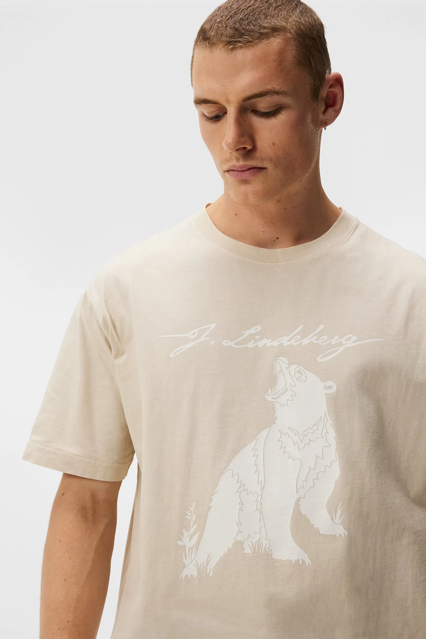 Ein Model trägt das Walsh Boxy Print Tee in Oyster Gray mit einem weißen Bären-Print und J.Lindeberg Schriftzug.