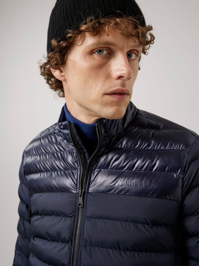 Mann mit Mütze trägt blaue, wattierte Rick Light Jacket mit Reißverschluss und kleinem J.Lindeberg Schriftzug.