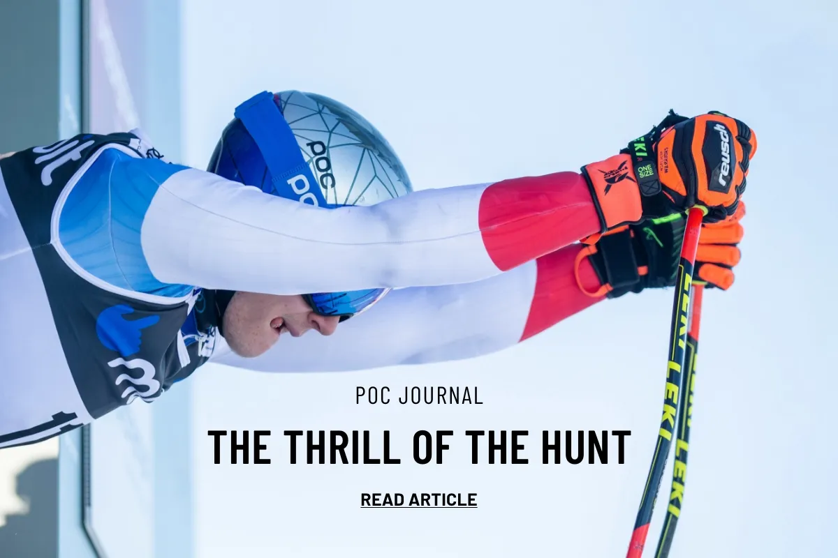 A ski racer with helmet and ski goggles holds his ski poles in front of a bright background.