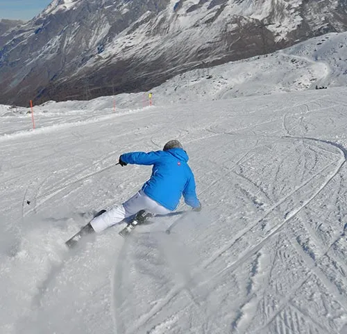 Sciatore con giacca blu e pantaloni bianchi su una pista innevata, sullo sfondo le montagne innevate.