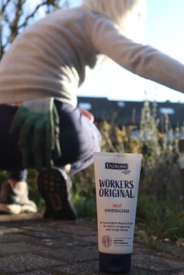 Un tube de baume pour les mains se trouve au premier plan, à l'arrière-plan une personne portant des gants de jardinage est agenouillée dans le jardin.