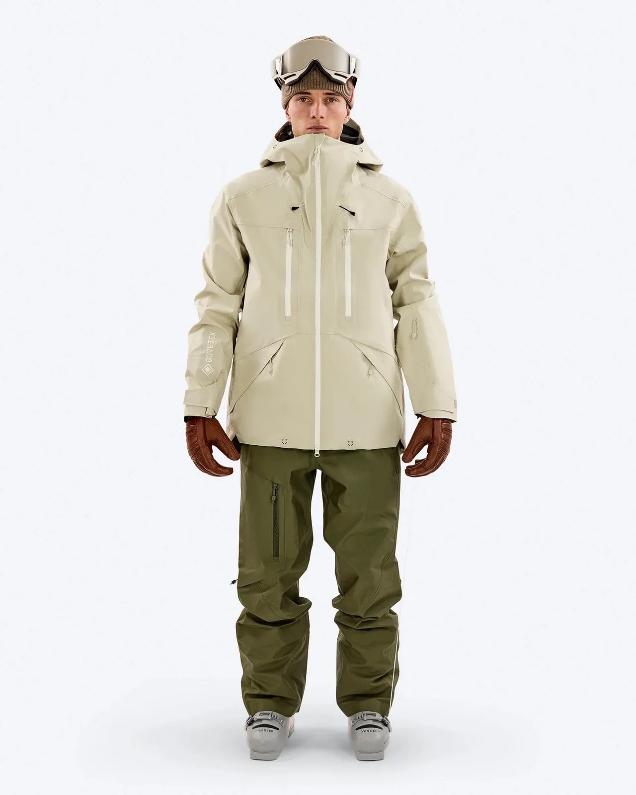A person in ski clothing stands in front of a white background. She is wearing a beige jacket, green trousers, brown gloves and ski goggles.