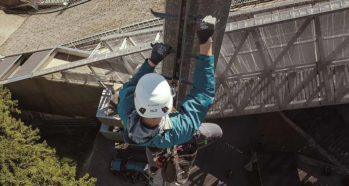 A climber in work clothes and helmet climbs up a ladder on a tall building. View from above.