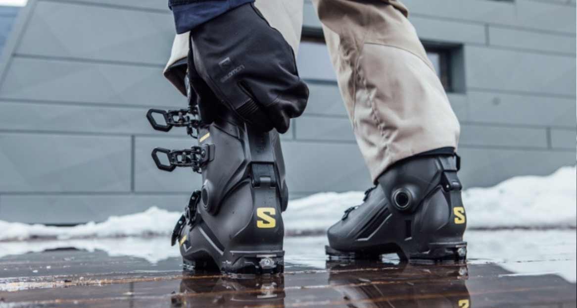 Zwei schwarze Skischuhe mit gelbem "S"-Logo auf glattem, reflektierendem Untergrund, teilweise mit Schnee. Eine Hand mit Handschuh schließt den linken Schuh.
