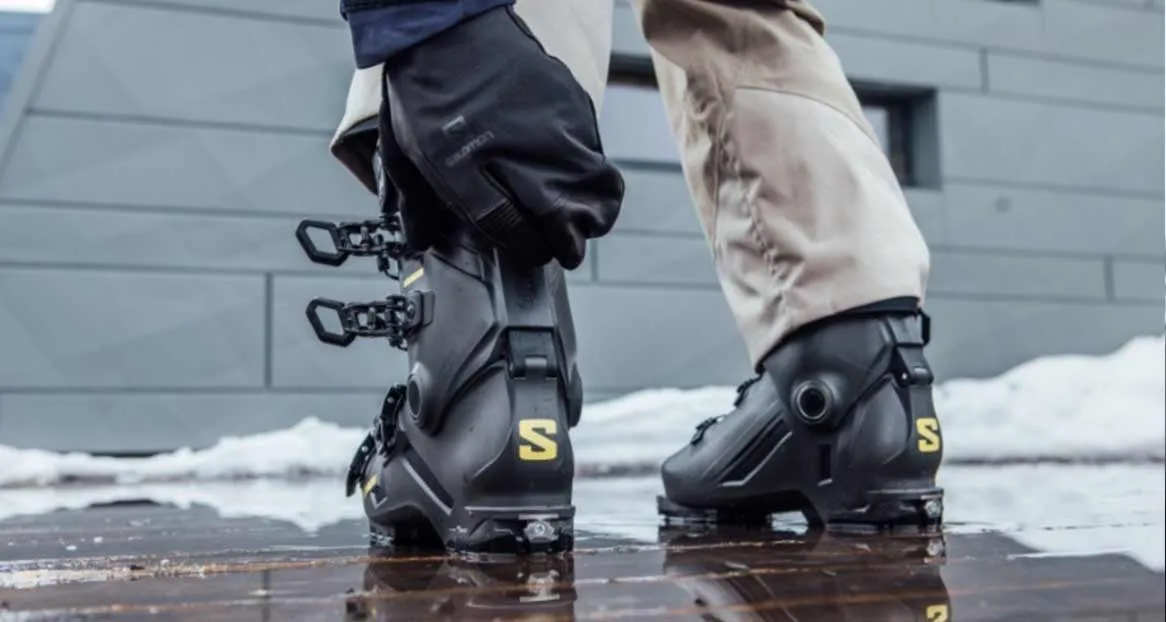 Zwei schwarze Skischuhe mit gelbem "S"-Logo auf glattem, reflektierendem Untergrund, teilweise mit Schnee. Eine Hand mit Handschuh schließt den linken Schuh.