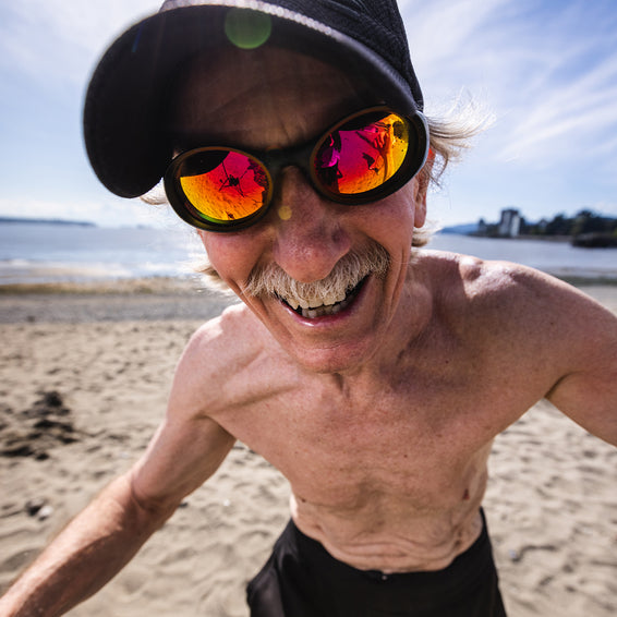 Lachender Mann mit Schnurrbart und reflektierender Brille am Strand, im Stil von The Slammer.
