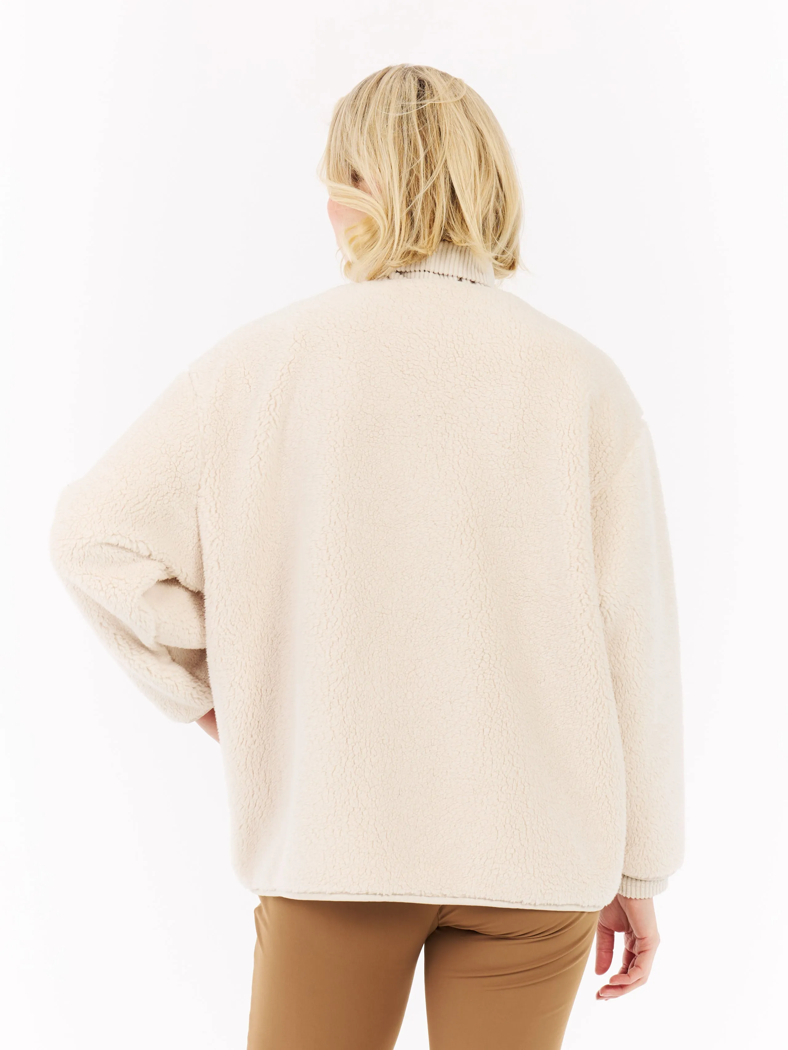 Rear view of a woman with blonde hair wearing a cream-colored teddy fur jacket and brown trousers against a white background.