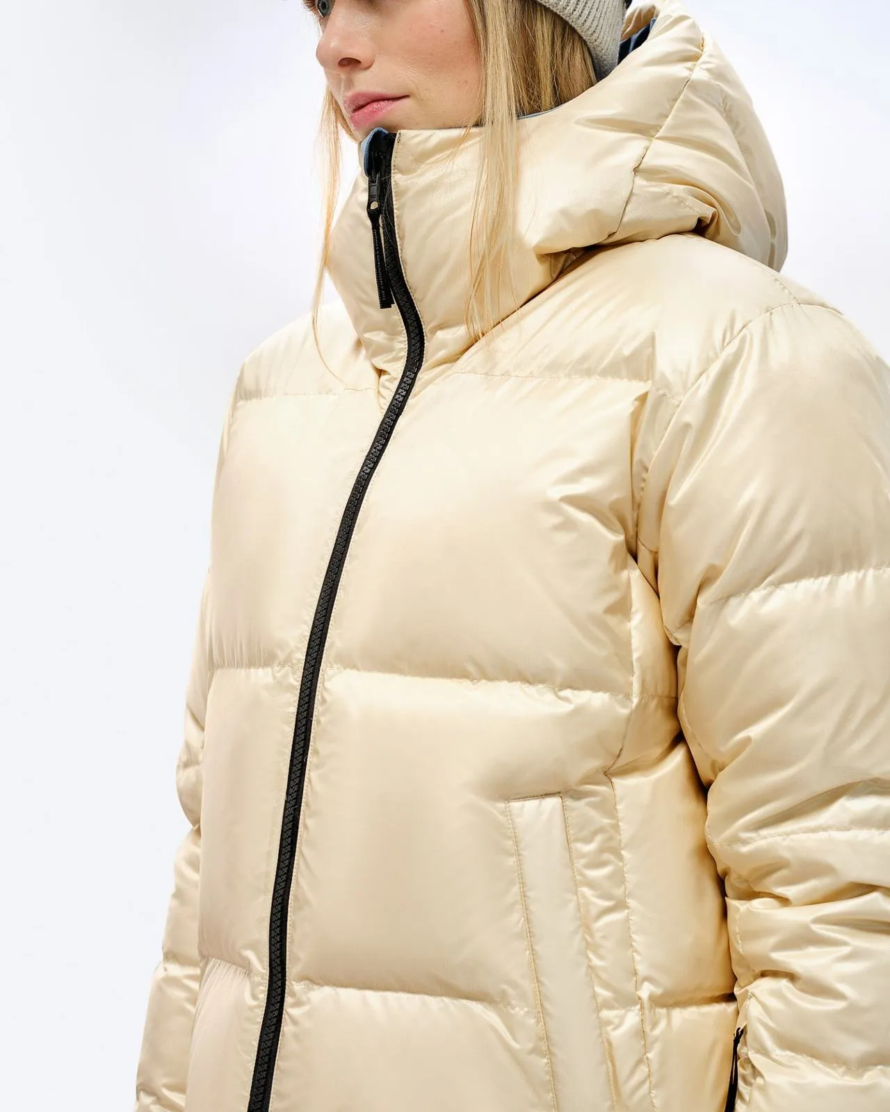 Person in shiny, beige down jacket with hood and black zipper. Quilted jacket, side view.