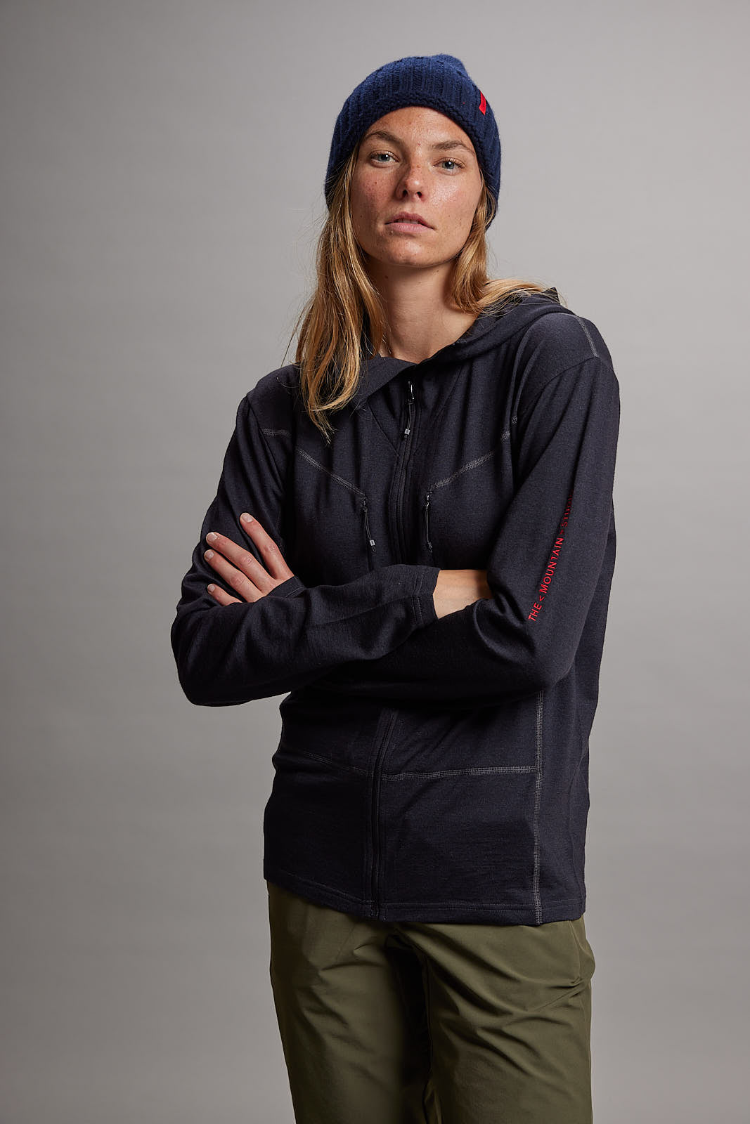 Eine Frau in dunkler Nuyarn Hood Jacket mit Kapuze und Reißverschluss. Sie trägt eine blaue Mütze und grüne Hose. Arme sind gekreuzt.