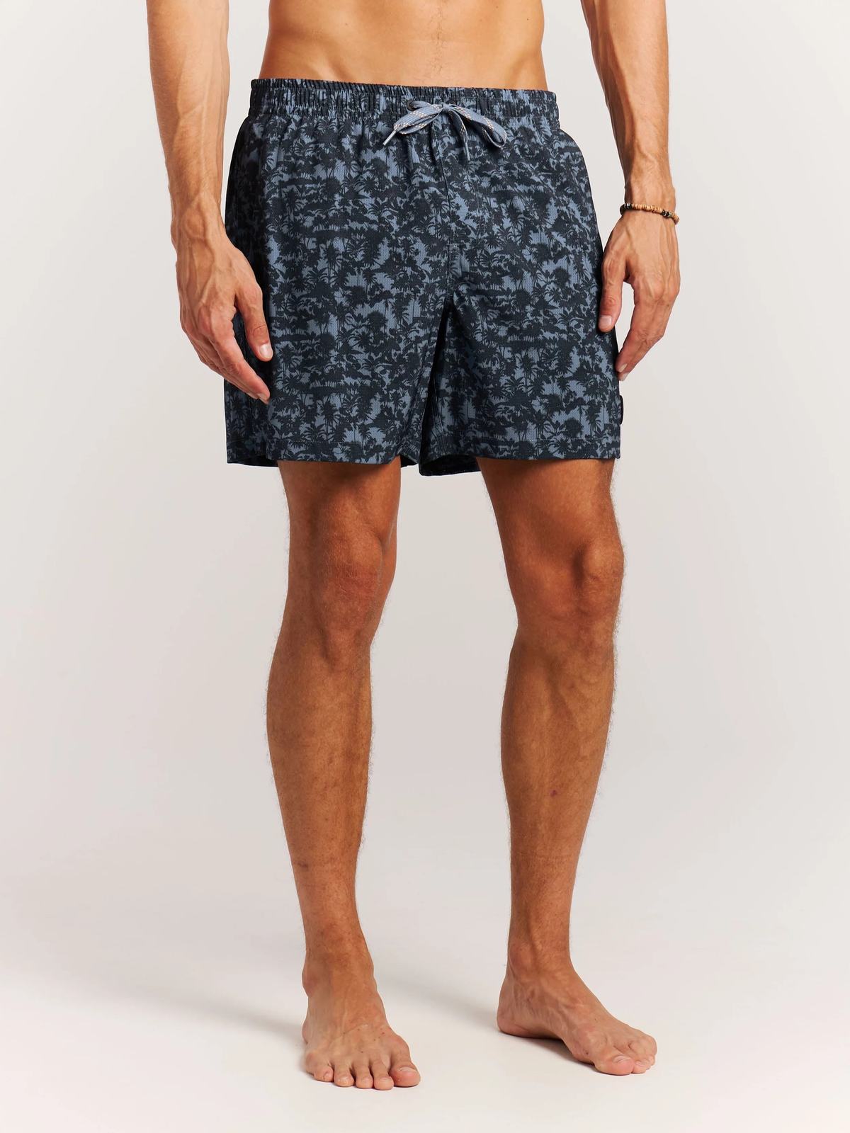 Ein Mann trägt Badeshorts mit Palmenmuster, stehend vor einem weißen Hintergrund.
