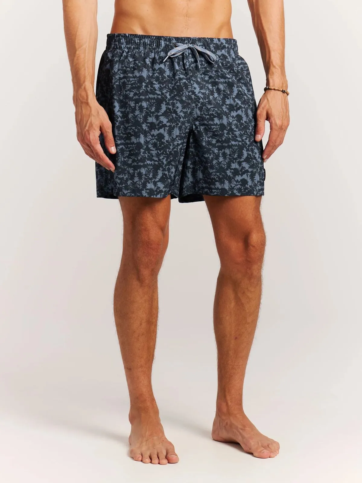 A man wears swimming shorts with a palm tree pattern, standing in front of a white background.