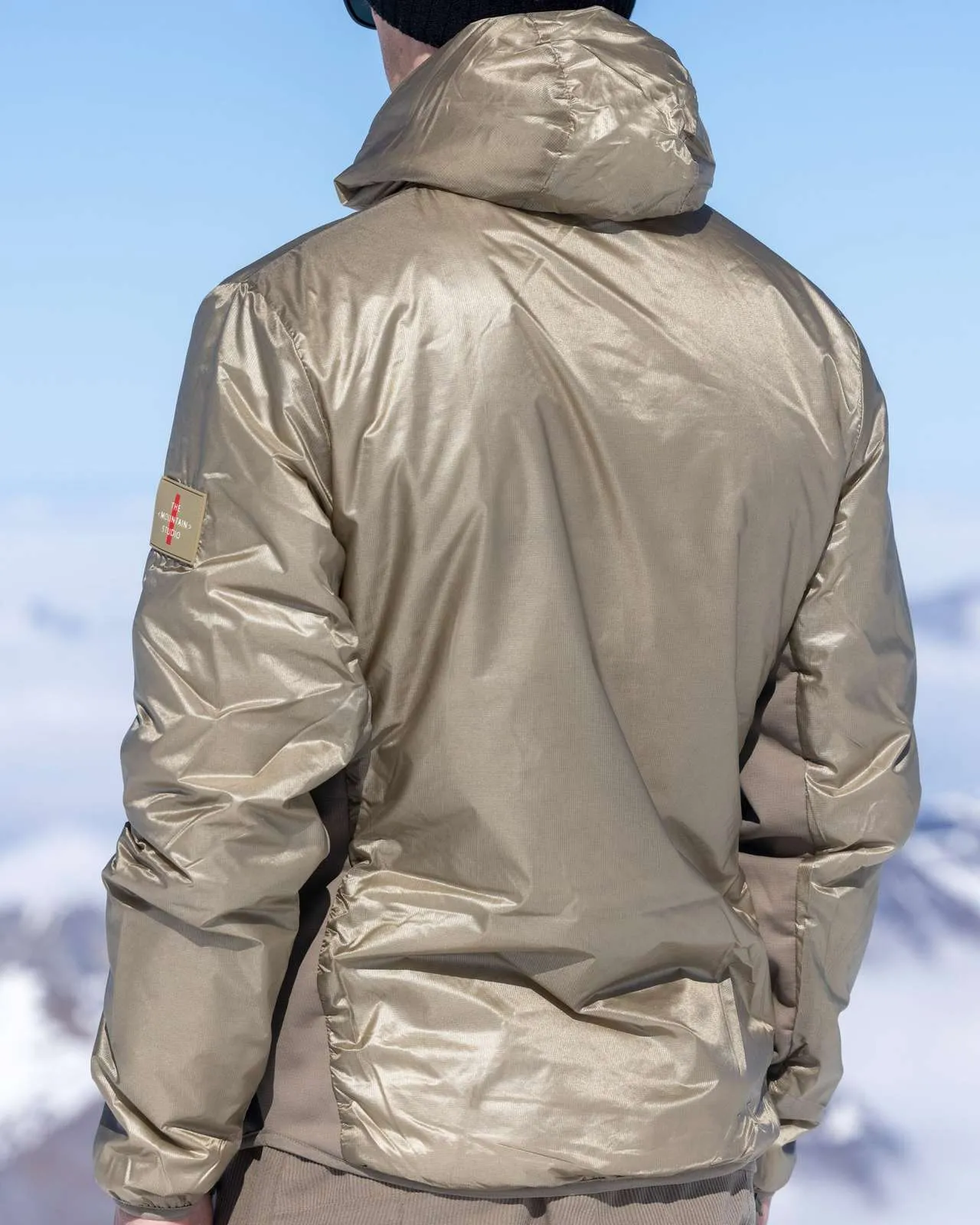 Back view of a person wearing a golden down jacket against a snowy mountain backdrop and blue sky.