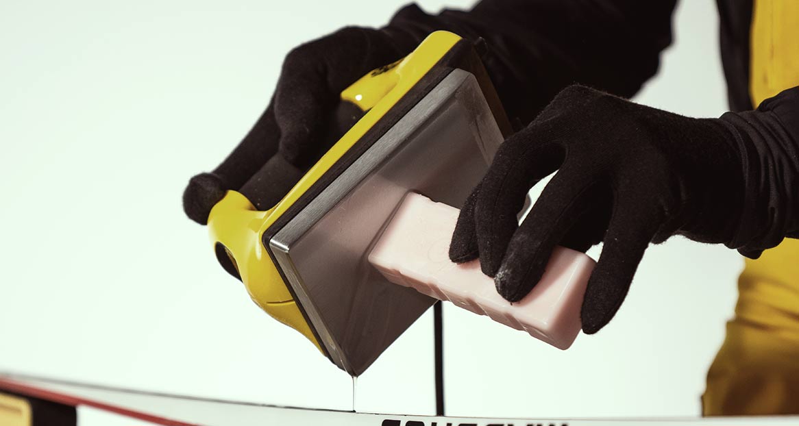 Eine Person in Handschuhen trägt Wachs mit einem Bügeleisen auf Skier auf.