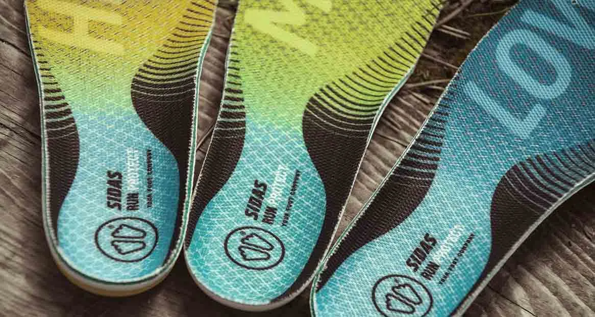Three colored shoe insoles from the brand Sidas lie on wood. Yellow, green and blue run in color and have black stripes.