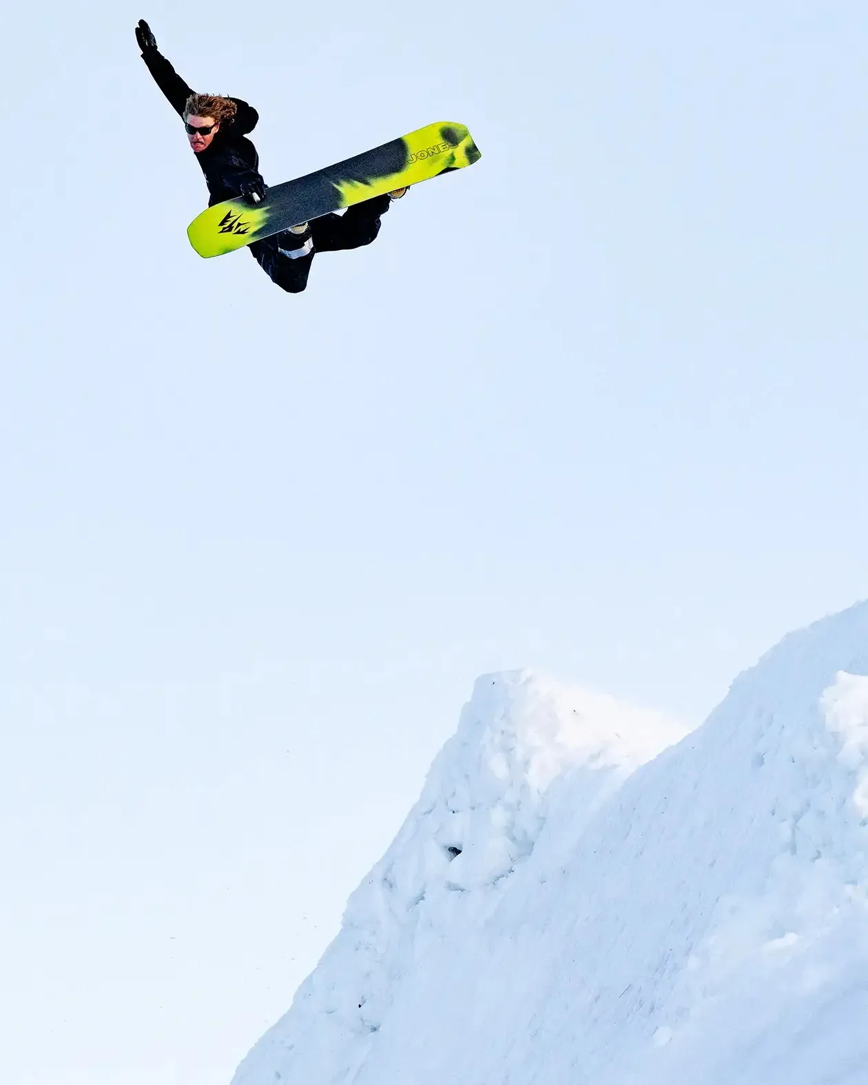 Snowboarder in volo davanti a un cielo blu e a una pista innevata. La persona indossa abiti neri e occhiali da sole.