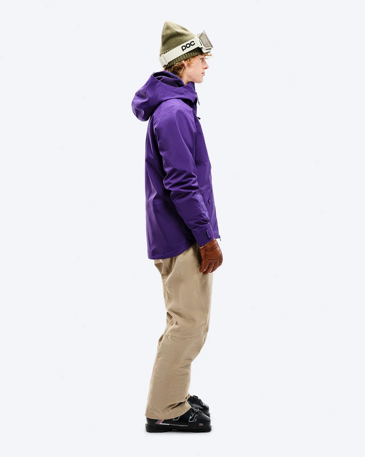 Person from the side in purple Z-2 GORE-TEX jacket, beige pants, ski boots, cap and goggles against a white background.