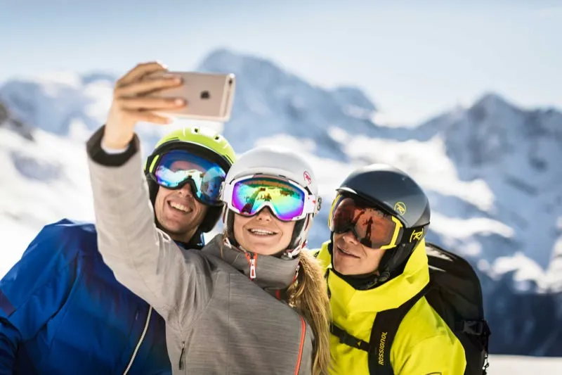 Tre sciatori con casco e occhiali si fanno un selfie davanti a montagne innevate.