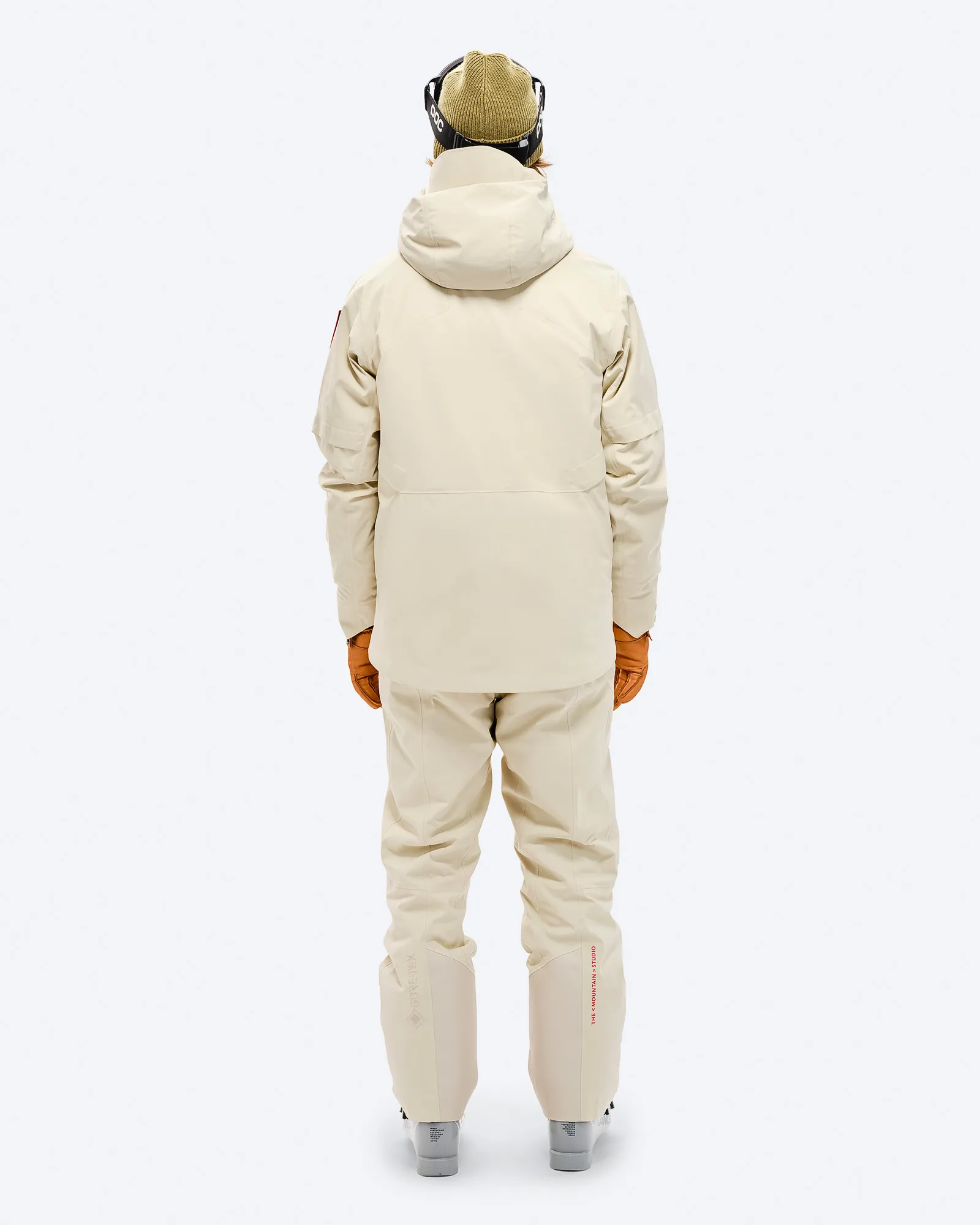 Person from behind in beige S-1 GORE-TEX 2L Stretch Insulated jacket and matching pants, brown gloves, ski goggles and cap.