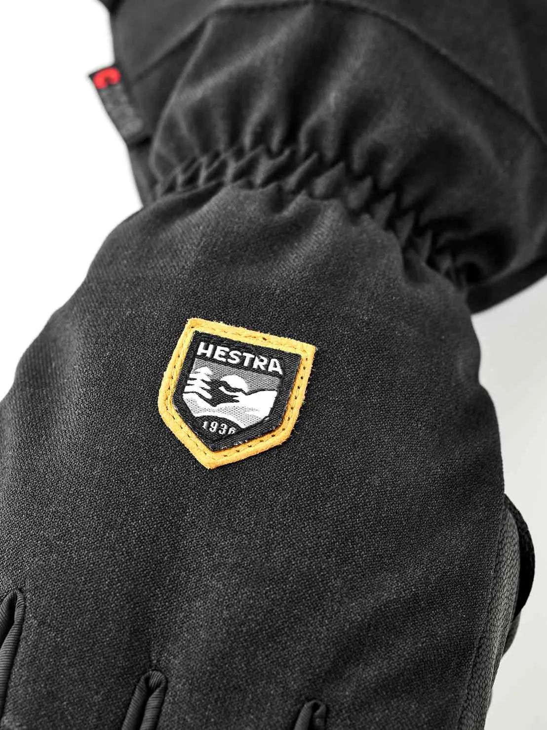 Detailansicht eines schwarzen Handschuhs mit Hestra-Logo, das Berge und das Gründungsjahr 1936 zeigt.