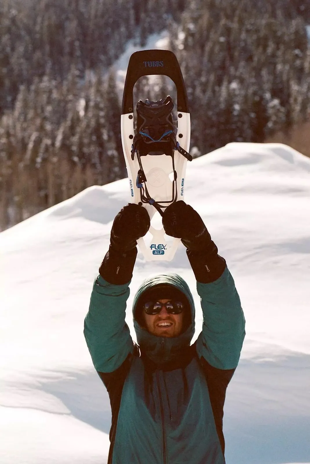 Man holding up Tubbs Flex Alp 25 snowshoe in white/blue with snowy mountain landscape in the background.