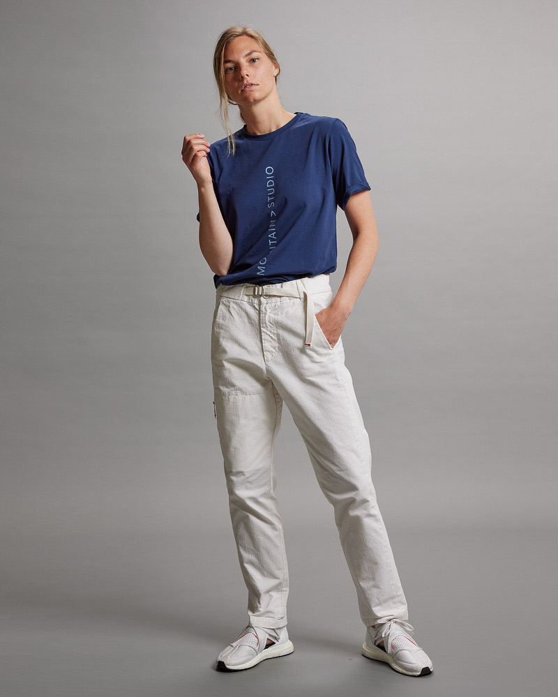 La femme porte un T-shirt bleu foncé avec une impression et un pantalon clair, debout sur un fond gris.