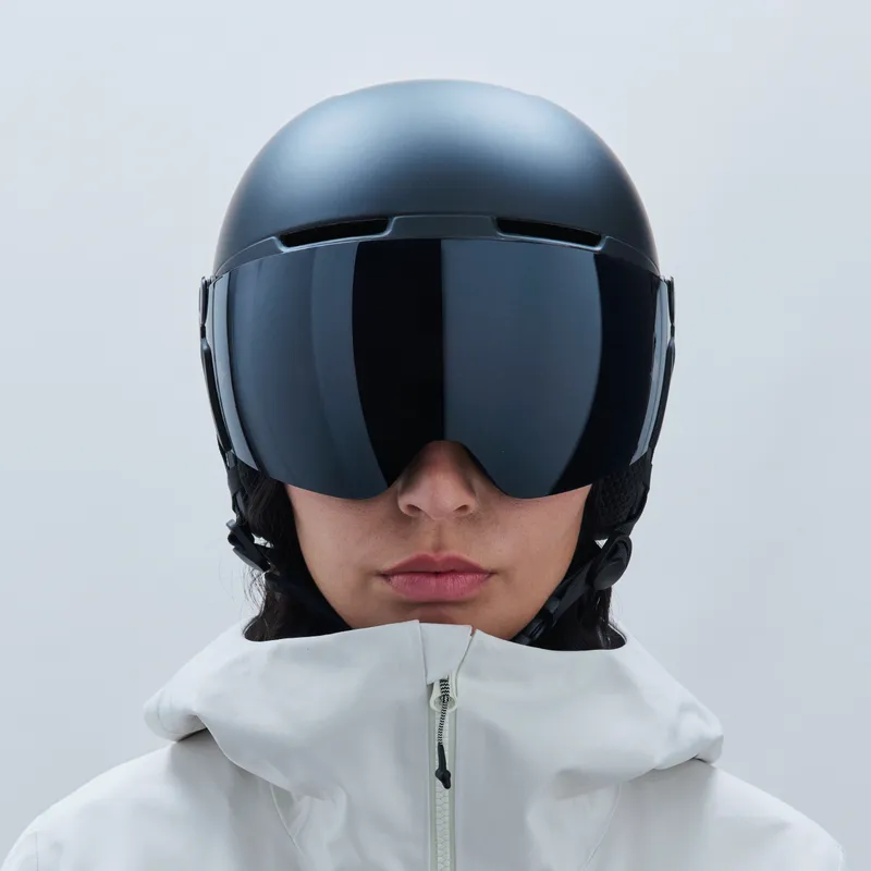 Una persona indossa un casco da sci nero con visiera. La visiera è oscurata. La persona indossa una giacca bianca con cerniera.