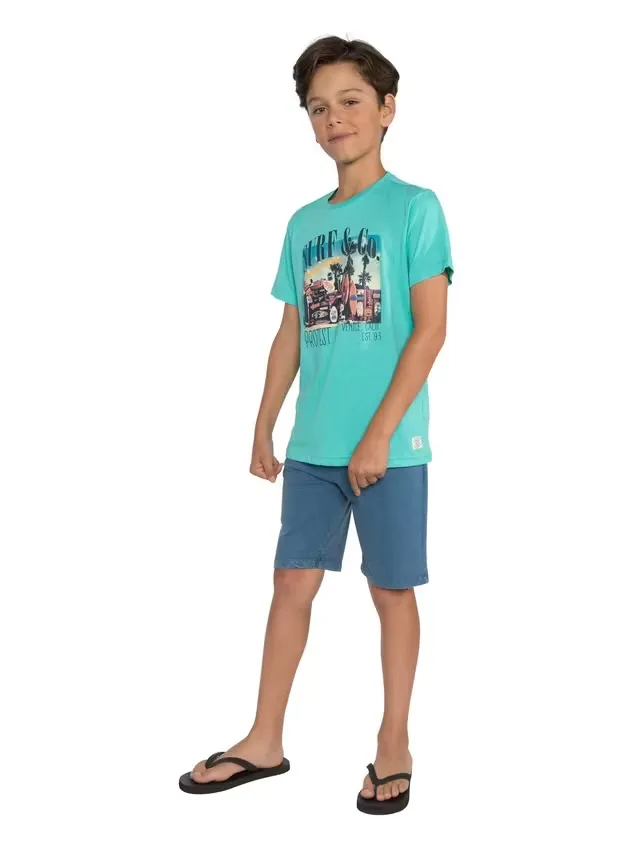 A boy is standing barefoot, wearing a turquoise T-shirt with a print of a car, palm trees, and the words "Surf & Co.," as well as blue shorts. He has brown hair and is wearing black flip-flops.