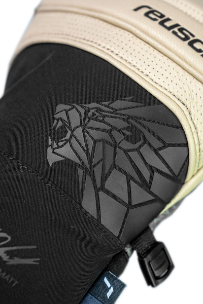 Nahaufnahme eines schwarzen Handschuhs mit geometrischem Löwenkopf-Design und beigem Lederteil mit Reusch-Logo.
