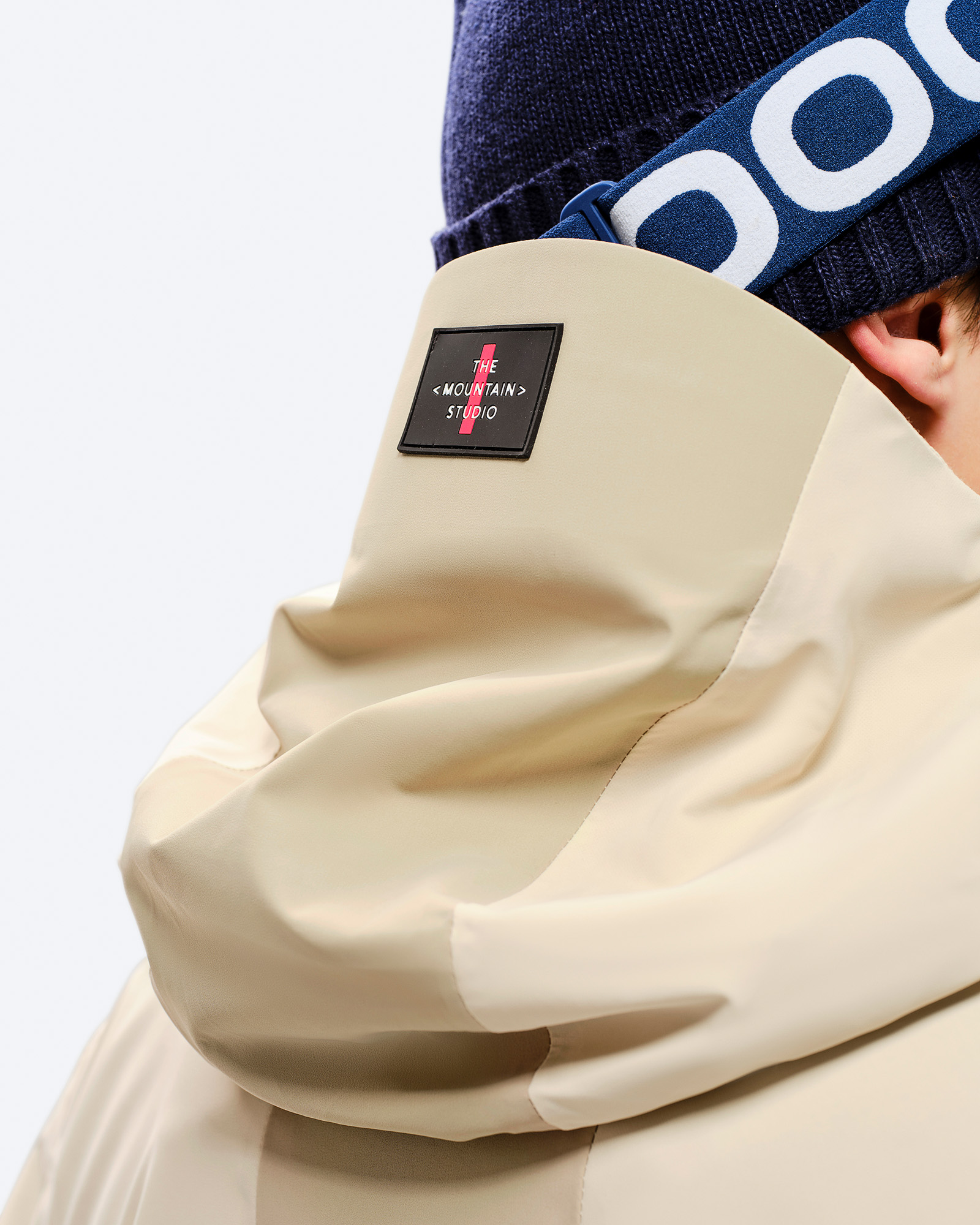 Beige Z-5 3L Jacke Nahaufnahme der Kapuze einer beigen Z-5 3L Hybrid Shell Jacke, mit Mütze und Skibrille im Hintergrund.