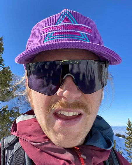 Person mit lila Kappe und dunkler, rechteckiger "The Cosmos Photchromic 2000" Sonnenbrille vor verschneiter Landschaft.