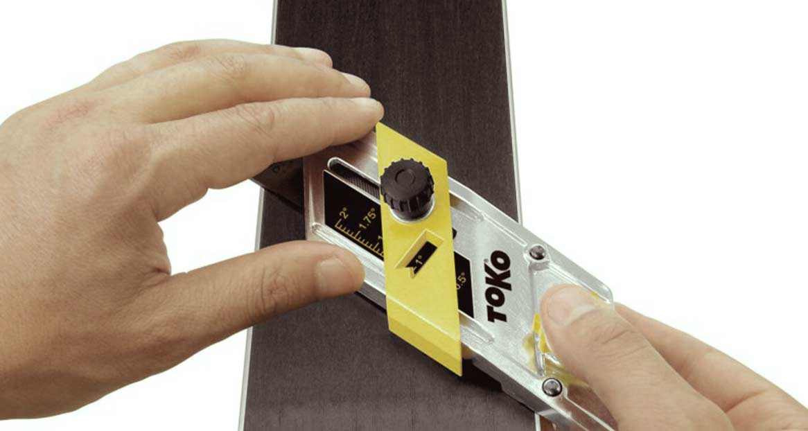 Toko Skiwerkzeug Anwendung Eine Hand führt ein Werkzeug der Marke Toko an der Kante eines Skis entlang. Das Werkzeug hat gelbe und silberne Elemente.