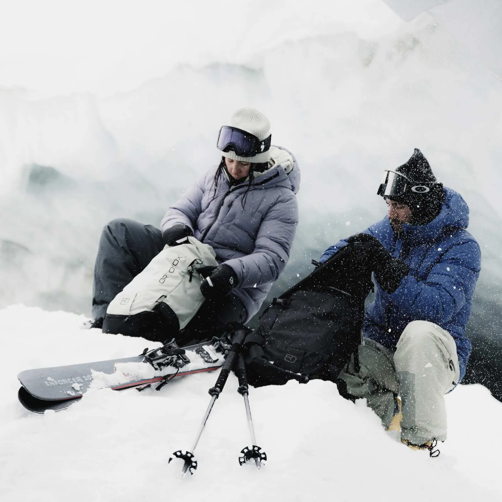 Zwei Skifahrer sitzen im Schnee vor einem Gletscher und öffnen ihre Rucksäcke. Skier und Stöcke liegen im Schnee.