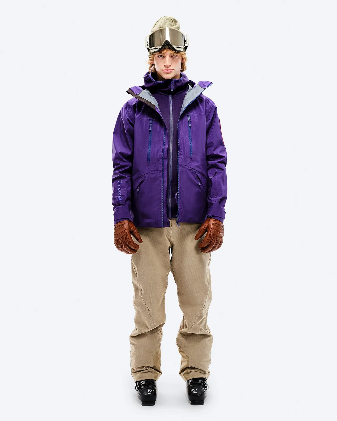 Man in purple GORE-TEX EPE 3L Shell Jacket, beige pants, ski boots and goggles, frontal on white.
