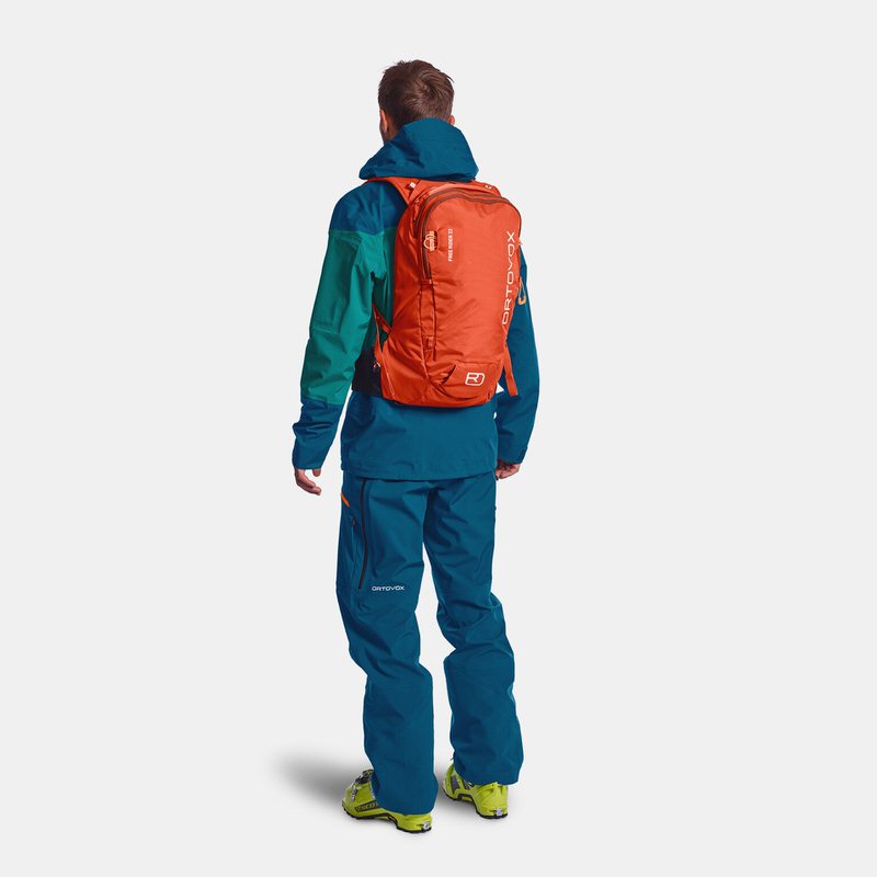 Mann von hinten mit orangefarbenem Ortovox Freerider 22 Rucksack. Blaue Jacke und Hose, gelb-grüne Schuhe.