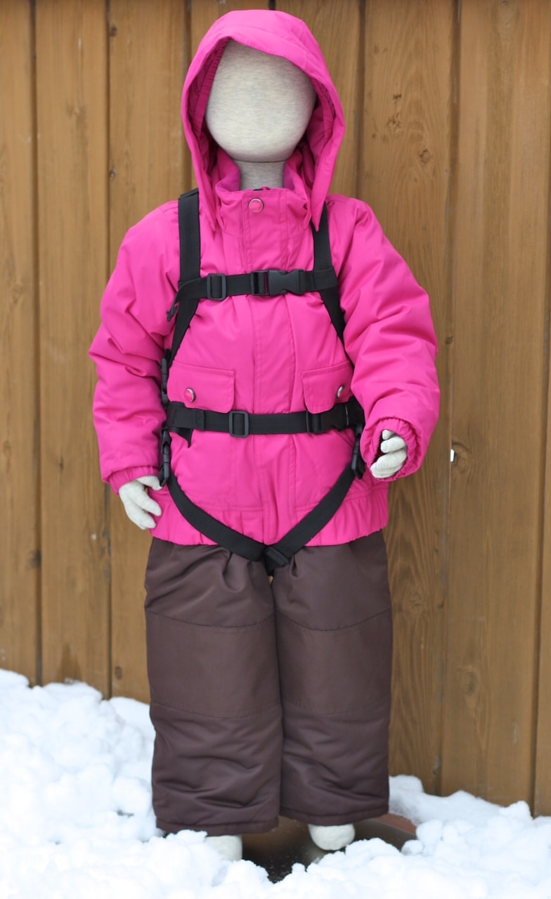 Poupée avec combinaison de ski rose et Lil' Ripper Gripper Aide au ski avec harnais noir devant un mur en bois.