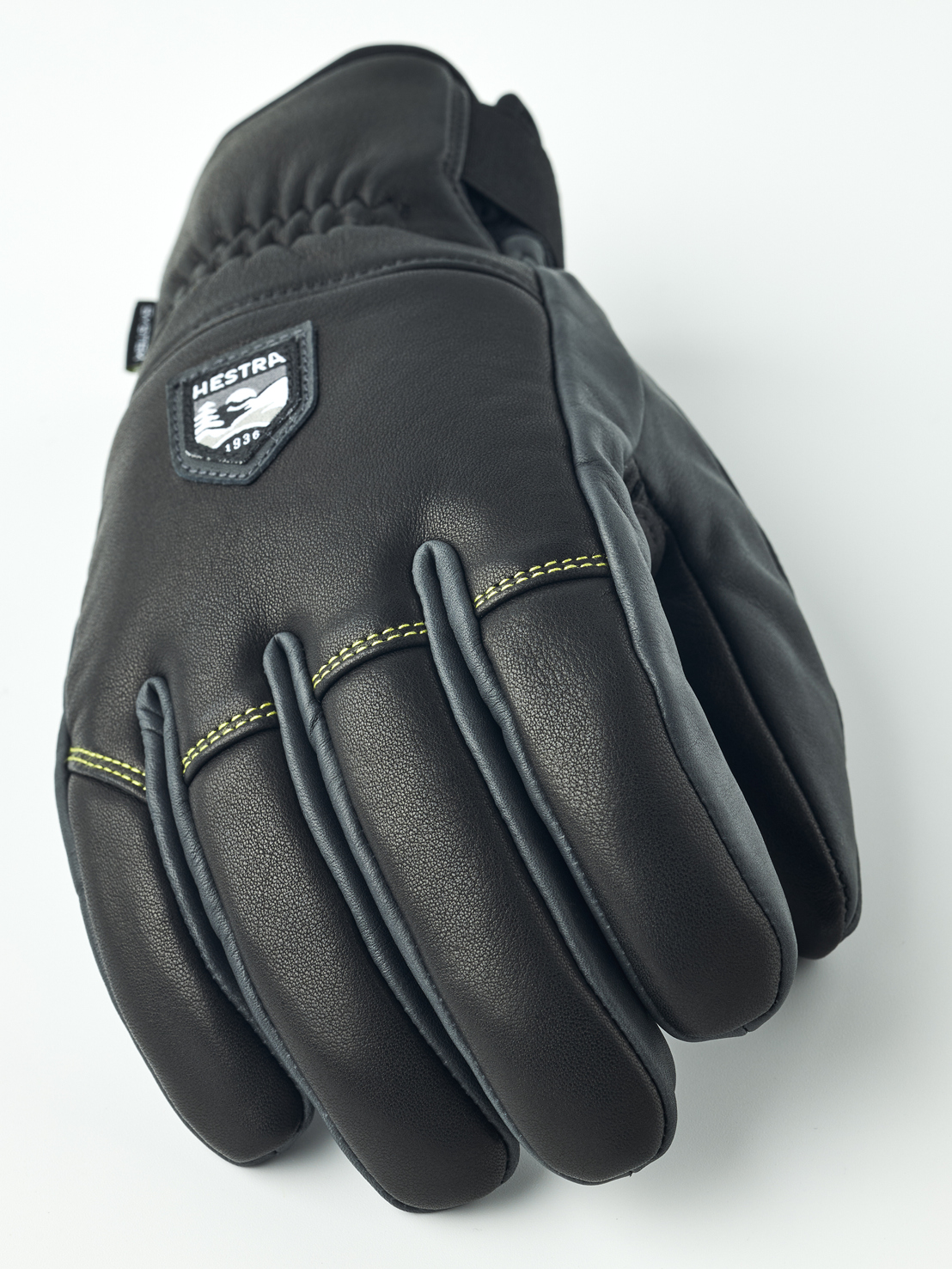 Eine schwarze Hestra Lederhandschuhe, frontal auf einem weißen Hintergrund fotografiert. Die Nähte sind gelb und Details grau.