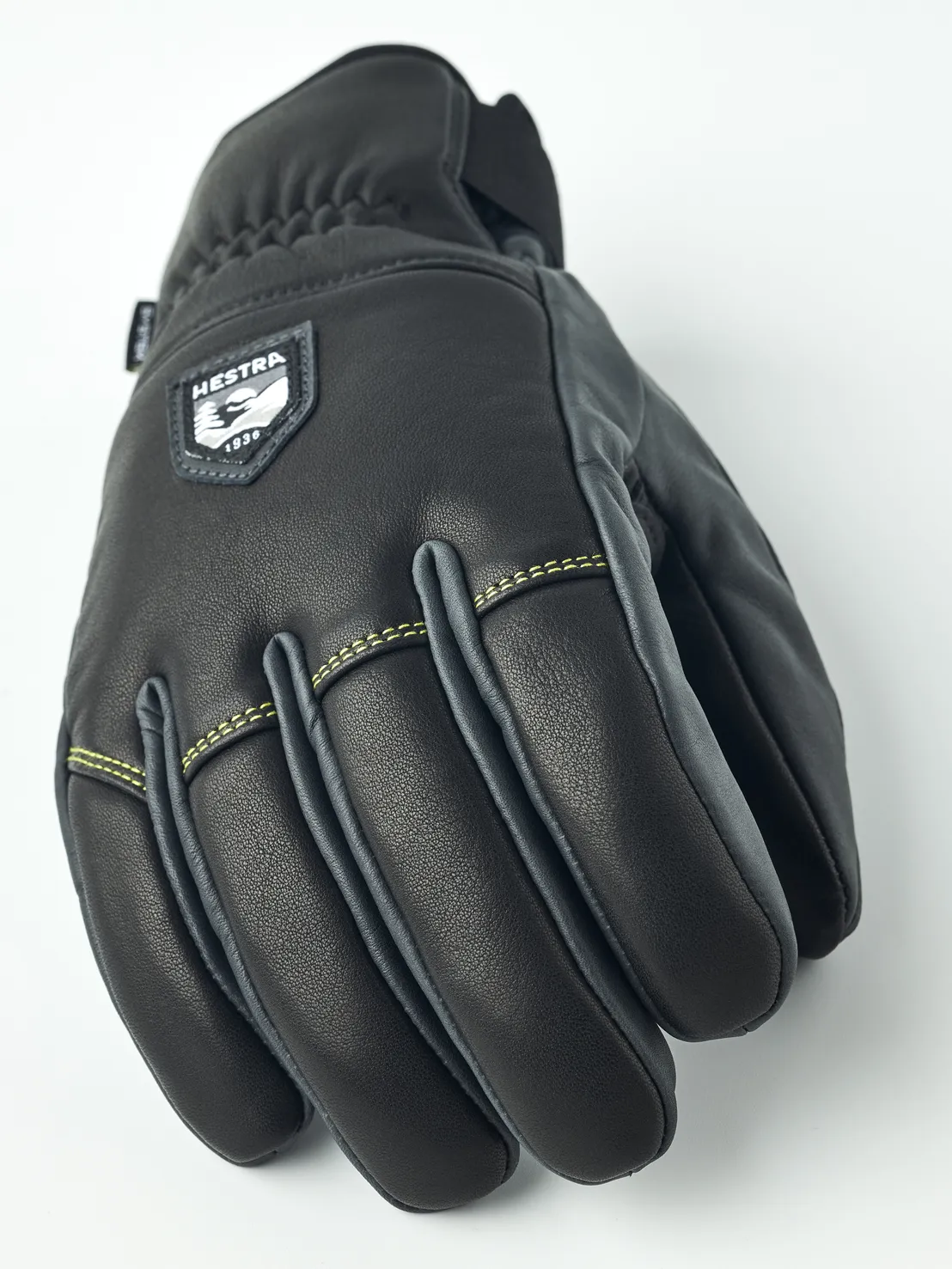 Eine schwarze Hestra Lederhandschuhe, frontal auf einem weißen Hintergrund fotografiert. Die Nähte sind gelb und Details grau.